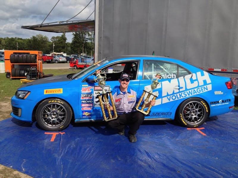 Driver kneeling beside a blue Volkswagen Jetta race car holding two trophies