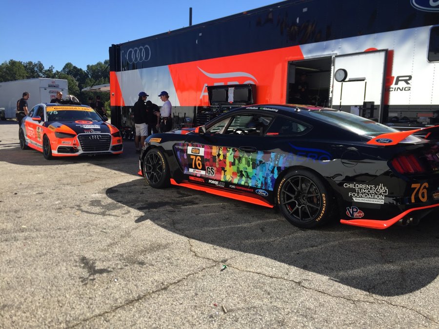 Audi RS3 and a tie-dye liveried Mustang GT4 number 76 parked behind the team hauler