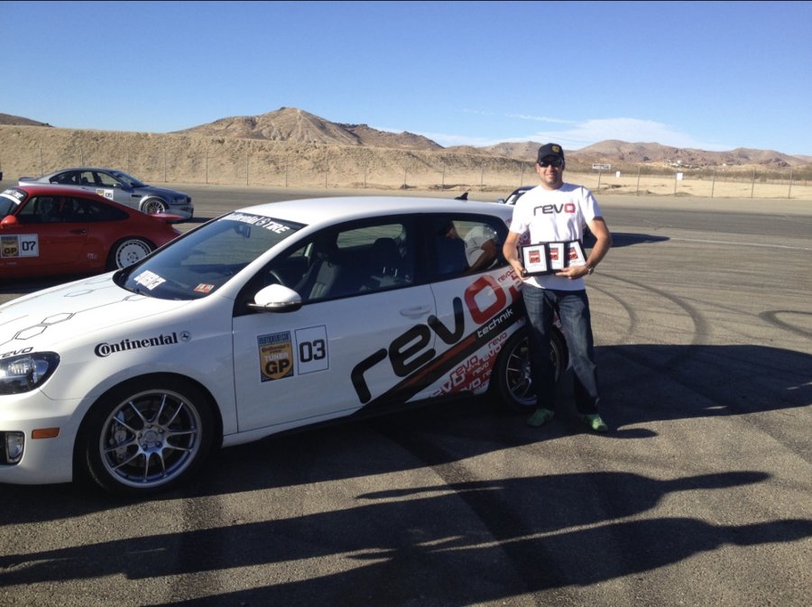 Driver holding award plaques beside a white Volkswagen at a desert airstrip event