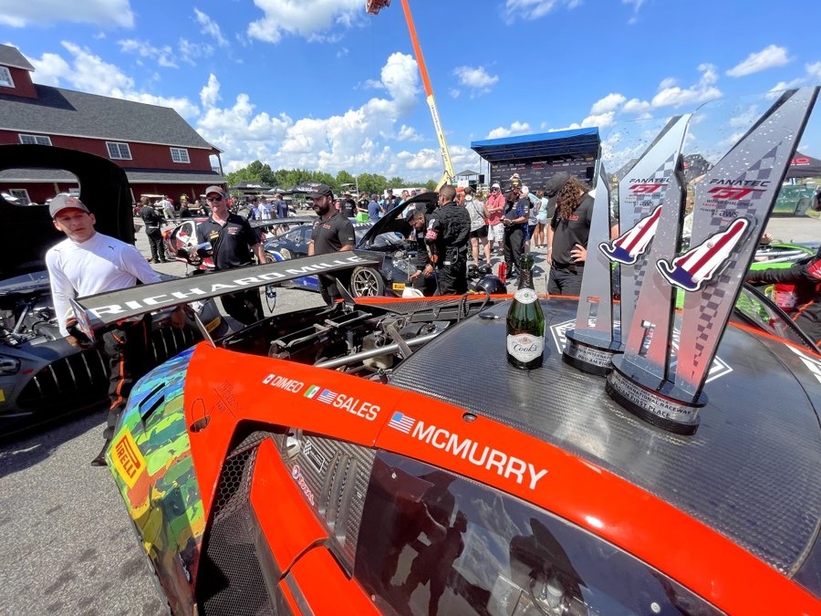 Trophies and champagne resting on the rear wing of an orange GT3 car in the paddock
